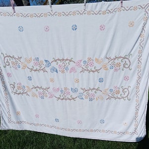 Könnte beinhalten: Weiße Tischdecke mit gesticktem Blumendesign in Rosa-, Blau-, Gelb- und Brauntönen. Das Design zeigt stilisierte Blumen und Schmetterlinge.