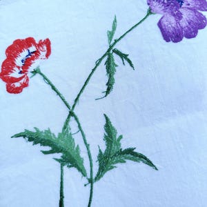 May include: Embroidered design of two flowers on a white fabric background. One flower is red and the other is purple. The flowers are connected by green stems and leaves.