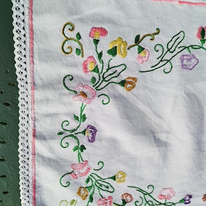 May include: White rectangular embroidered cloth with a floral design in pink, yellow, purple, and green. The edges are trimmed with a pink border and a white lace trim. The cloth is likely a decorative item.