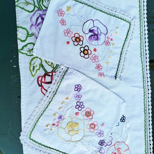 Peut inclure: Tissu blanc avec des motifs floraux brodés dans des tons de rose, violet, jaune et vert. Le tissu est bordé d'une dentelle délicate.