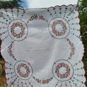 Può includere: Un panno quadrato in tessuto bianco con bordi smerlati rifiniti con filo rosso. Il centro è decorato con quattro ghirlande floreali circolari ricamate nei toni del rosa, arancione e verde, con steli radianti che si estendono verso gli angoli.