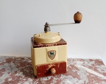 May include: A vintage, hand-crank coffee grinder made of metal and wood. The grinder is cream-colored with brown accents and features a drawer for collecting the ground coffee. The grinder has a label that reads "Peugeot Paris" and depicts a lion.