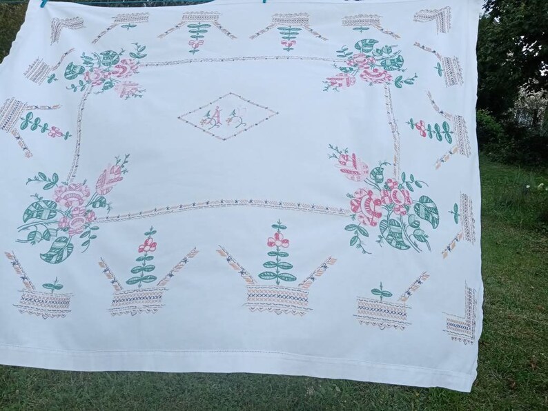 May include: White tablecloth with embroidered floral designs in pink, green, and brown. The design features repeating patterns of flowers, leaves, and baskets.