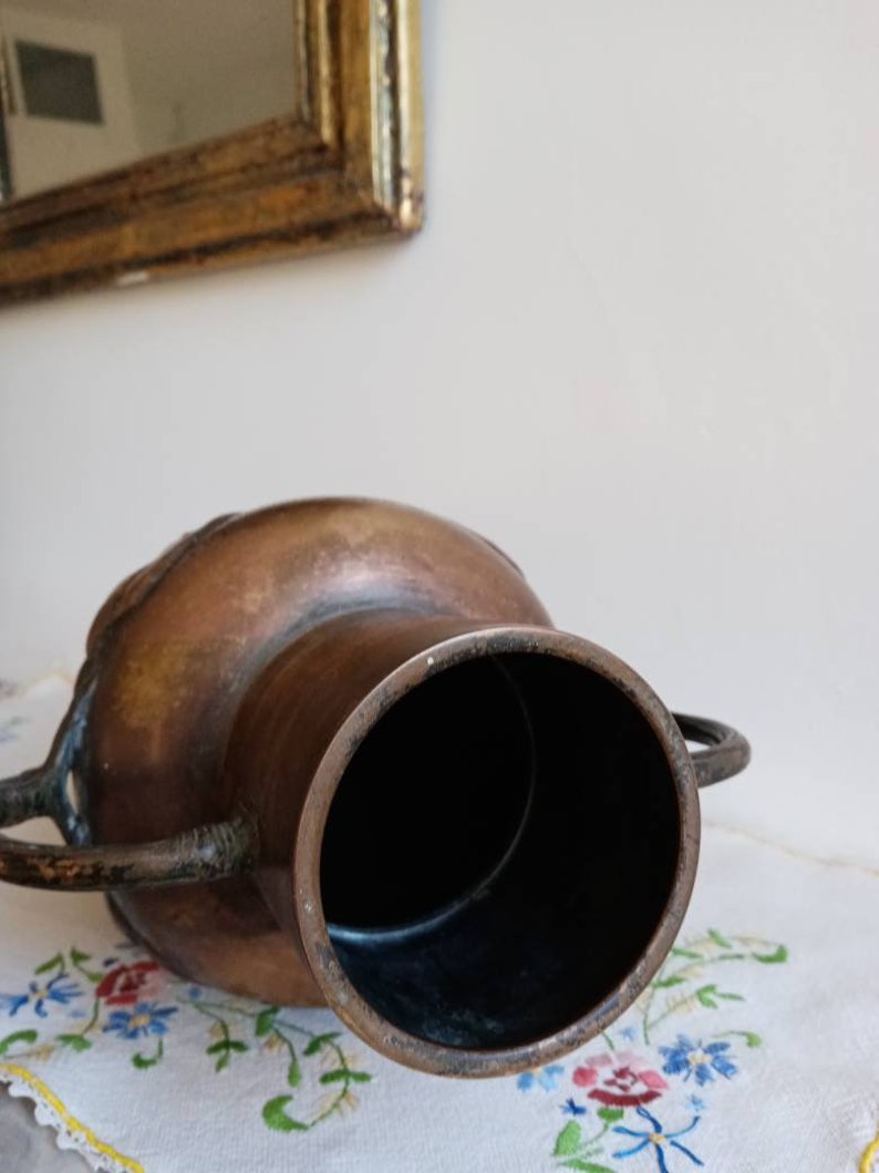 Op de afbeelding: Een grote, koperkleurige vaas met twee handvatten. De vaas heeft een donkerbruin interieur en een versleten, verweerde uitstraling.