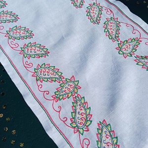 May include: White linen table runner with a red, green, and yellow paisley pattern embroidered along the edges.