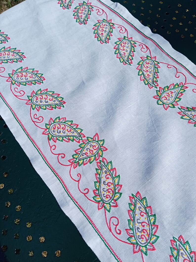 May include: White linen table runner with a red, green, and yellow paisley pattern embroidered along the edges.