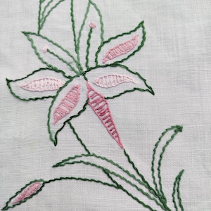 May include: A close-up of a white linen fabric with a pink and green embroidered flower design. The flower has five petals and is surrounded by leaves.