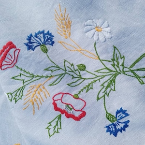Op de afbeelding: Wit linnen stof met geborduurde bloemen in rood, blauw, wit en geel. De bloemen omvatten klaprozen, korenbloemen en een madeliefje.