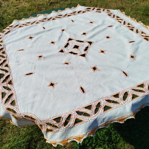 Può includere: Tovaglia bianca con un intricato bordo in pizzo ricamato in marrone e arancione. Il centro della tovaglia presenta un grande disegno quadrato con forme di diamante più piccole.