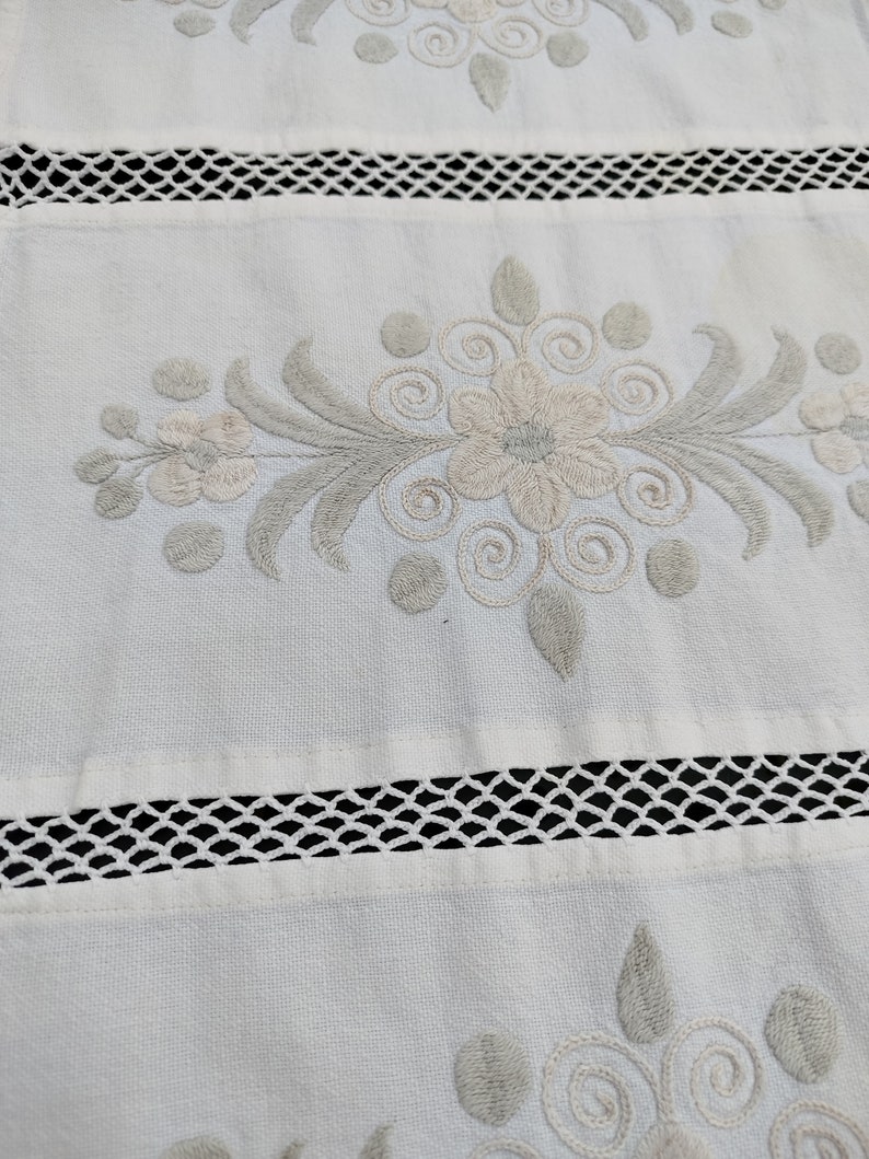 May include: White fabric with a delicate embroidered floral design in shades of gray and white. The fabric has a lace trim along the bottom edge.
