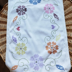 May include: White table runner with embroidered flowers in shades of blue, pink, purple, orange, and green. The flowers are arranged in a circular pattern around the border of the runner.