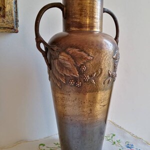 Op de afbeelding: Een hoge, antieke koperen vaas met twee handvatten en een reli&euml;fdesign van bladeren en bessen. De vaas heeft een donkerbruine patina en een gouden bovenkant.