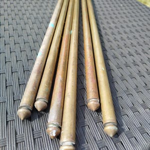 Peut inclure: Un ensemble de cinq tiges cylindriques longues en laiton avec une finition métallique patinée. Chaque tige présente une extrémité en forme de cône pointu. Les tiges sont disposées parallèlement et affichent une couleur or terne.