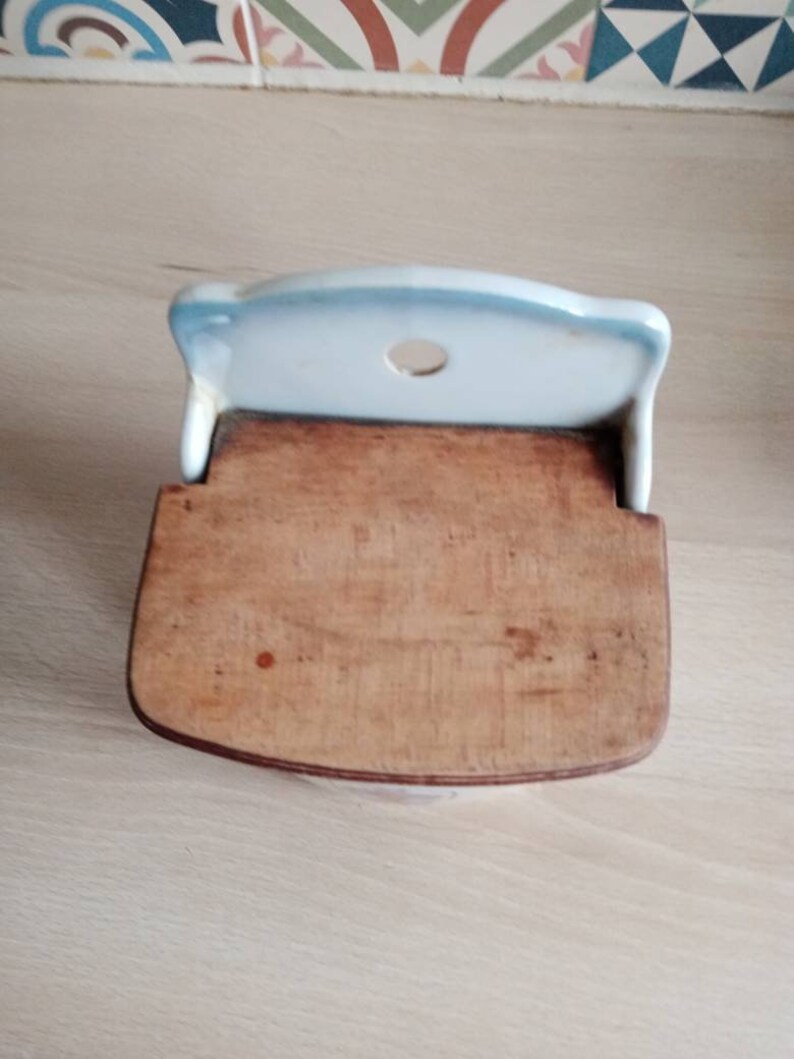 May include: A white ceramic wall-mounted spice rack with a wooden lid. The rack has a round hole in the top and a blue and white trim.