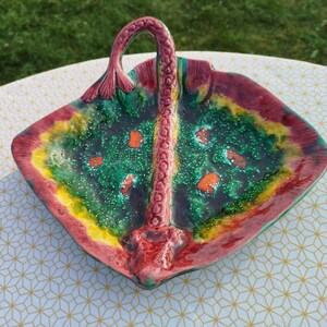 May include: A colourful ceramic serving dish shaped like a stingray with a handle. The dish is decorated with green, red, yellow and orange glaze and has a speckled finish.