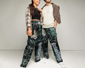 May include: Two people wearing matching black sweatpants with a green and white pattern and the text "Brutino Design" in white. The sweatpants have a drawstring waist and are paired with white sneakers. One person wears a black top and plaid shirt, the other a white shirt and brown jacket.