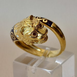 Könnte beinhalten: Ein goldener Ring mit einem detaillierten Löwenkopf-Design. Die Augen des Löwen sind mit roten Edelsteinen besetzt. Die Ringschiene ist mit schwarzem Emaille und goldenen Schnörkeln verziert.
