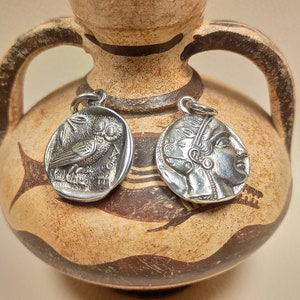 May include: Two silver pendants with detailed designs. One pendant features an owl, the other a profile of a person. The pendants are displayed against a backdrop of a brown and beige ceramic vase, suggesting a historical or classical theme.