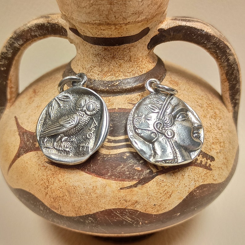 May include: Two silver pendants with detailed designs. One pendant features an owl, the other a profile of a person. The pendants are displayed against a backdrop of a brown and beige ceramic vase, suggesting a historical or classical theme.