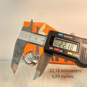May include: A silver-colored pendant with a profile of a person's face is measured with a digital caliper. The caliper displays a measurement of 0.89 inches. The pendant has a small loop at the top for a chain.
