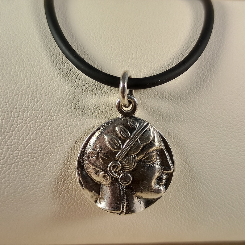May include: A silver-colored pendant necklace featuring a detailed profile of a person with a laurel wreath, suspended from a black rubber cord. The pendant has an antique coin design, showcasing classical artistry.