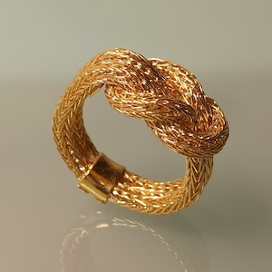 May include: A gold-toned ring with a braided knot design. The ring is made of woven metal and has a polished finish.
