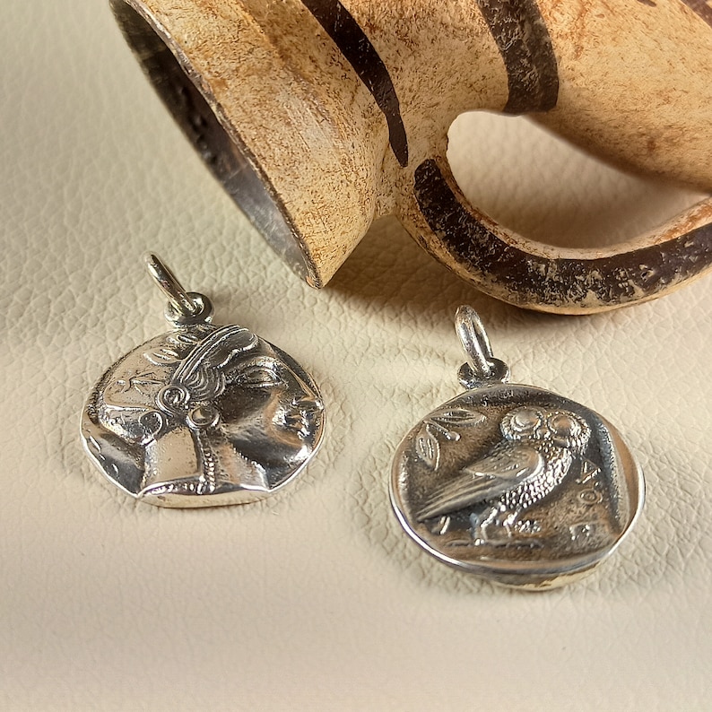 May include: Two silver-colored coin pendants. One pendant features a profile of a woman's face, the other an owl with an olive branch. The pendants are round with a loop at the top for a chain. A tan ceramic jug is in the background.