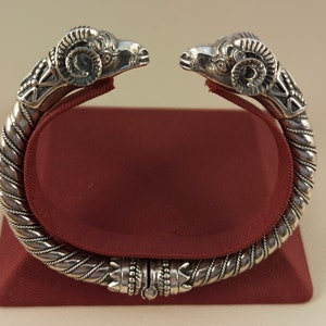 Puede incluir: Brazalete plateado con extremos de cabeza de carnero. El brazalete presenta cabezas de carnero detalladas en cada extremo, con intrincados diseños a lo largo de la banda. Se exhibe sobre un soporte de terciopelo rojo.