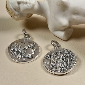 Puede incluir: Dos colgantes de plata, uno con el perfil de una mujer con casco y el otro con una figura alada sosteniendo un bastón. Ambos colgantes son redondos y tienen un diseño en relieve.