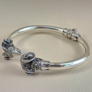 Puede incluir: Brazalete plateado con diseños de cabeza de carnero en cada extremo. Las cabezas de carnero tienen cuernos y rasgos faciales detallados. El brazalete tiene un diseño articulado y probablemente está hecho de plata de ley.