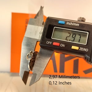 May include: A small, gold-colored pendant with an intricate design is measured with a digital caliper. The pendant measures 0.12 inches in diameter. The caliper displays the measurement in millimeters as 2.97 mm. The background is orange.