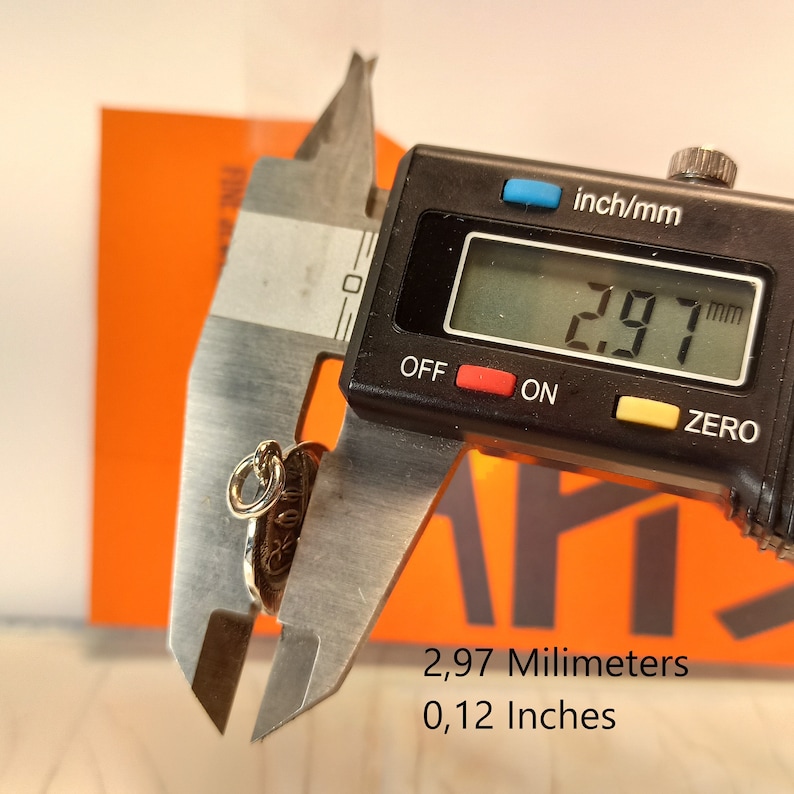 May include: A small, gold-colored pendant with an intricate design is measured with a digital caliper. The pendant measures 0.12 inches in diameter. The caliper displays the measurement in millimeters as 2.97 mm. The background is orange.
