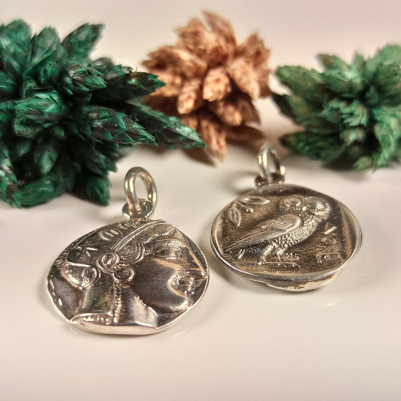 May include: Two silver-colored pendants with a loop for a chain. One pendant features a profile of a person, and the other depicts an owl. The pendants are on a white surface with green and brown decorative elements in the background.