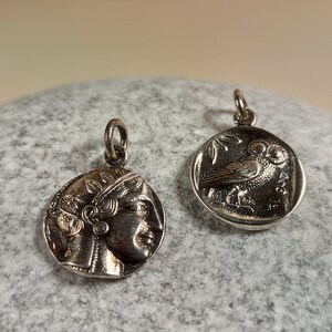 May include: Two silver-toned coin pendants. One pendant features a profile of a woman's face, the other depicts an owl perched on a branch. Both pendants have a small loop at the top for attaching to a chain or bracelet. The pendants are displayed on a gray, textured surface.