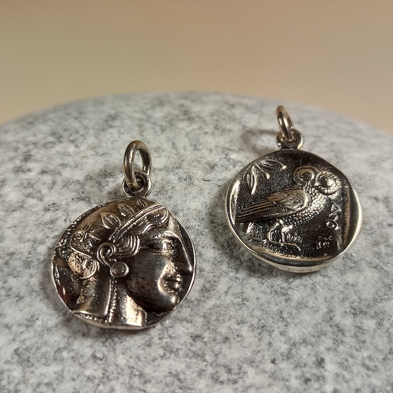 May include: Two silver-toned coin pendants. One pendant features a profile of a woman's face, the other depicts an owl perched on a branch. Both pendants have a small loop at the top for attaching to a chain or bracelet. The pendants are displayed on a gray, textured surface.