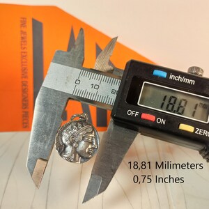 May include: A silver-colored pendant with a profile of a classical figure is measured with a digital caliper. The pendant is approximately 0.75 inches (18.8 mm) in diameter. The image includes text that reads "FINE JEWELS EXCLUSIVE DESIGNERS PIECES."