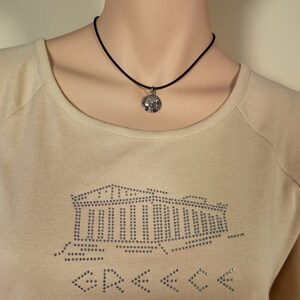 May include: A silver pendant necklace with a black cord. The pendant is round with a detailed design. The model is wearing a beige long-sleeve shirt with a rhinestone design of a building and arrows.