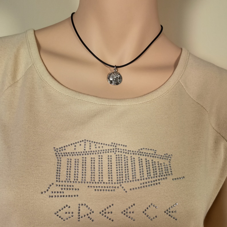 May include: A silver pendant necklace with a black cord. The pendant is round with a detailed design. The model is wearing a beige long-sleeve shirt with a rhinestone design of a building and arrows.