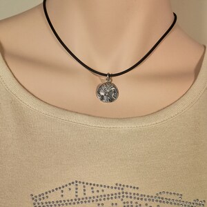 May include: A silver-toned pendant necklace featuring a detailed circular charm with a classical design. The charm is suspended from a thin, black cord. The necklace is displayed on a person wearing a light beige top.