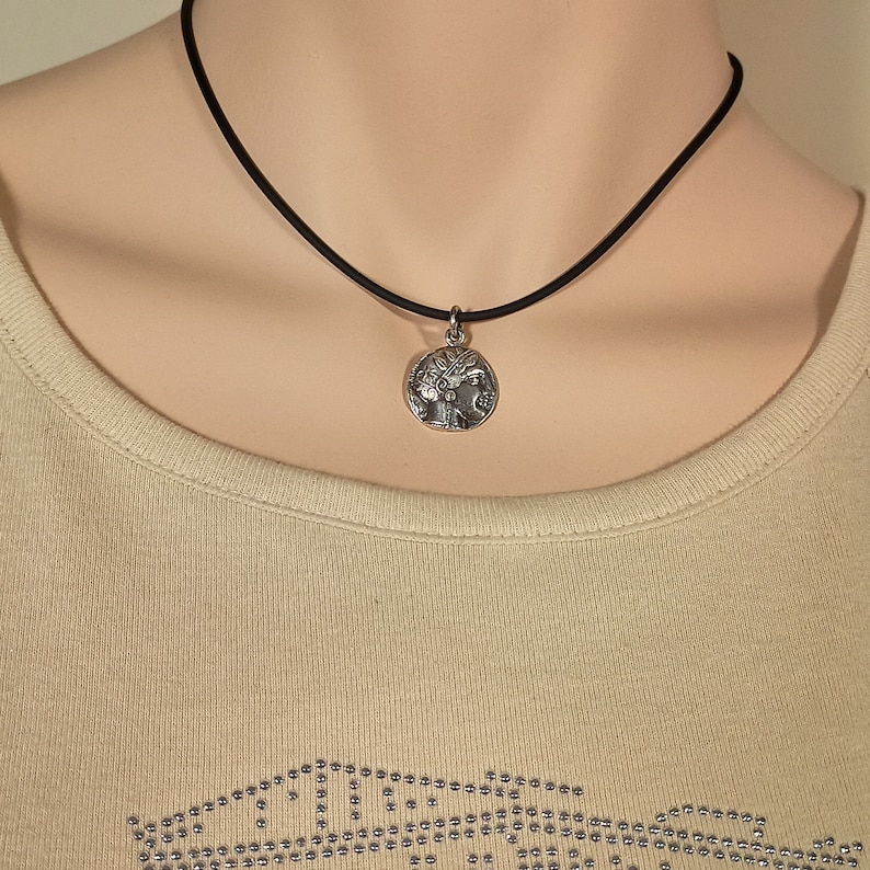 May include: A silver-toned pendant necklace featuring a detailed circular charm with a classical design. The charm is suspended from a thin, black cord. The necklace is displayed on a person wearing a light beige top.