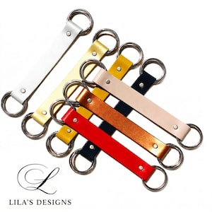 May include: A collection of leather keychains in various colors, including white, gold, yellow, tan, copper, navy, and red. Each keychain features a rectangular leather strap with a metal ring at each end. The brand name "Lila's Designs" is visible.