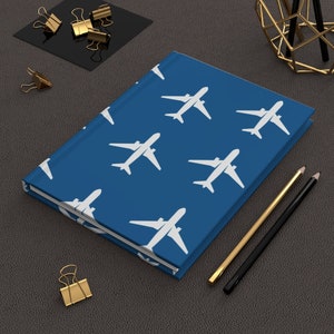 Puede incluir: Una libreta azul con un patrón de avión blanco. La libreta está sobre una superficie gris con dos lápices y clips de papel dorados.