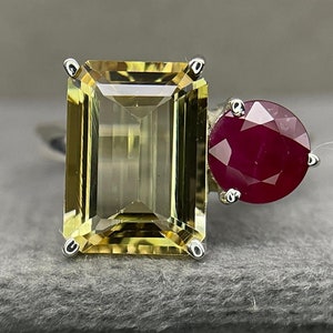 May include: A silver ring featuring a large, rectangular, light yellow gemstone and a smaller, round, deep red gemstone. The gemstones are set in a minimalist design, showcasing their natural beauty. The ring is displayed on a gray surface.