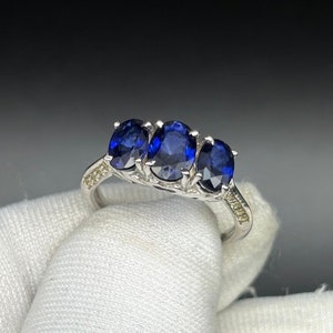 May include: A silver ring with three oval-shaped blue sapphires set in a row. The ring is accented with small white diamonds.