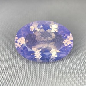 May include: An oval-cut gemstone with a pale lavender hue. The gem has a faceted surface that reflects light, creating a sparkling effect. The stone is set against a light gray background.
