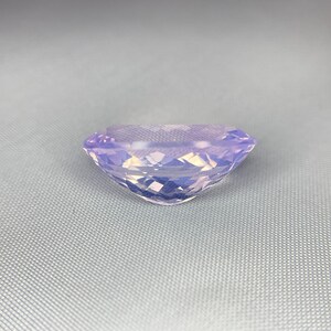 May include: A faceted, oval-shaped gemstone with a soft lavender hue. The gem is cut to maximize light reflection, creating a sparkling effect. The stone is set against a light gray background, highlighting its color and clarity. This gemstone is suitable for jewelry.