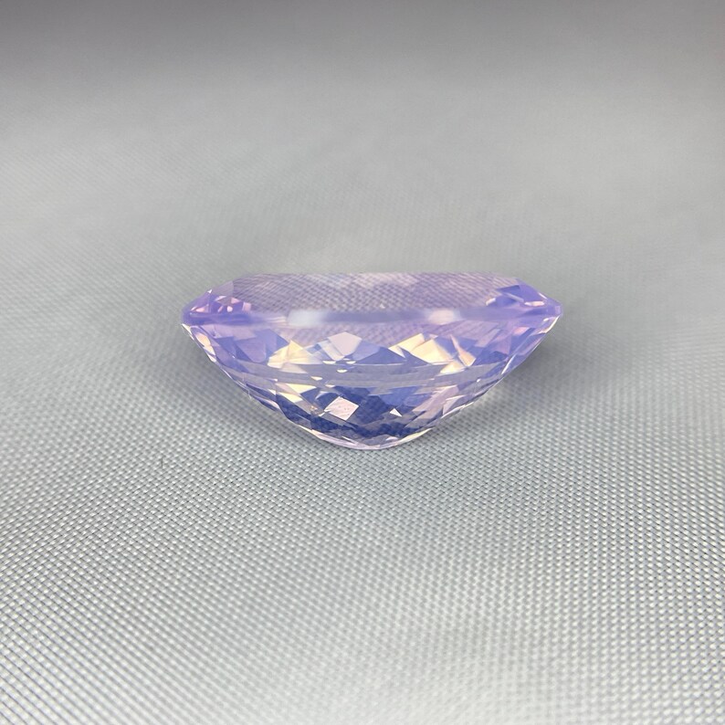 May include: A faceted, oval-shaped gemstone with a soft lavender hue. The gem is cut to maximize light reflection, creating a sparkling effect. The stone is set against a light gray background, highlighting its color and clarity. This gemstone is suitable for jewelry.