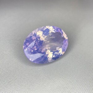 May include: An oval-shaped gemstone with a faceted surface. The gem has a soft, translucent appearance with a blend of lavender and pale yellow hues. The stone is set against a gray background.