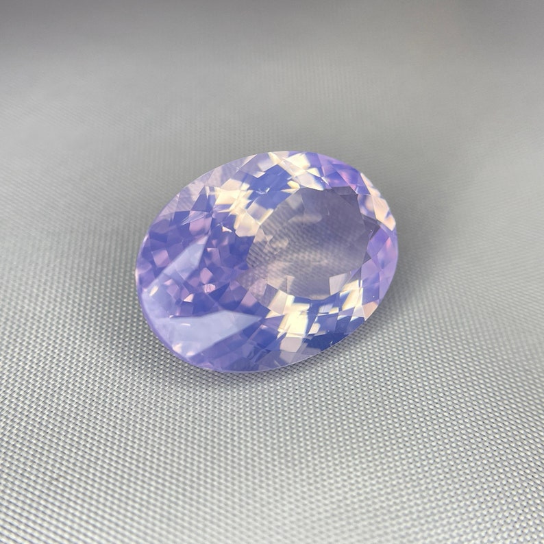 May include: An oval-shaped gemstone with a faceted surface. The gem has a soft, translucent appearance with a blend of lavender and pale yellow hues. The stone is set against a gray background.