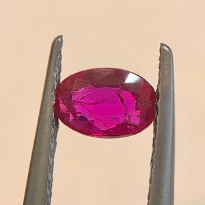 Puede incluir: Una gema de rubí ovalada y facetada, sujeta por unas pinzas plateadas. El rubí es de un color rojo-rosado intenso y vibrante, con múltiples facetas que capturan la luz. Las pinzas están enfocadas, resaltando la claridad de la gema.